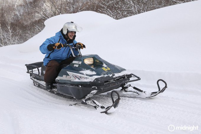 A person wearing a blue jacket and silver helmet riding a snowmobile (marked '129') through a snowy landscape, with the watermark 'midnight' in the bottom right corner.