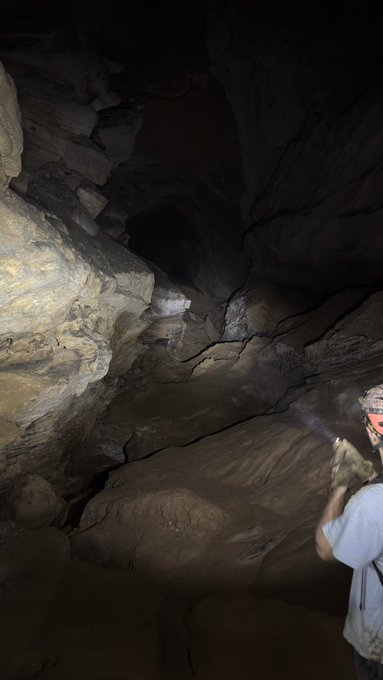 A person wearing a helmet explores a dimly lit cave with rocky formations, using a headlamp to illuminate the path.