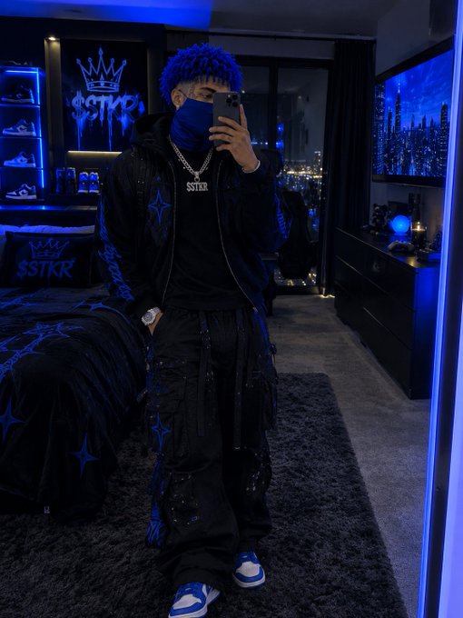 A man with blue hair takes a mirror selfie in a room with blue neon lighting; he wears a blue face covering, black jacket and pants with 'SSTKR' branding, and blue and white sneakers.