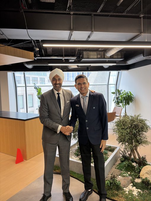 Two men in business suits smile and shake hands in a modern office space with exposed ceilings and indoor plants.