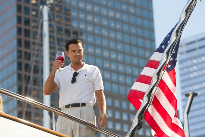 A man in a white polo shirt stands on a yacht holding a glass of red wine, with an American flag and tall city buildings in the background, projecting an image of luxury and success. The scene evokes the lifestyle of financial winners—fitting for a post challenging followers to explain how they'd turn $1,000 Solana into $2,000 through trading, gambling, or reselling.