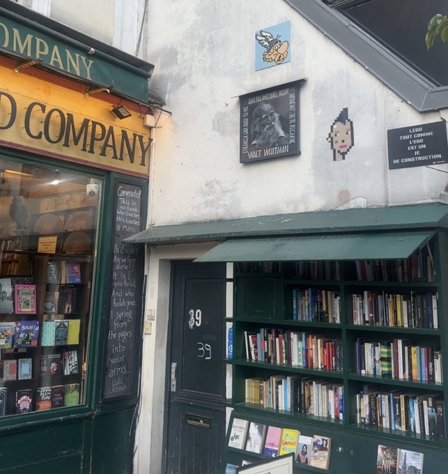 The exterior of a bookshop named 'COMPANY' with green trim, numbered 39, featuring outdoor bookshelves and a plaque for Walt Whitman.