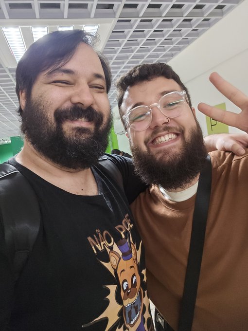 Two bearded men take a cheerful selfie together, with one wearing a black shirt featuring a giraffe graphic and the Portuguese text "NÃO P..." (likely "Não Pare"), while the other in a brown shirt and clear glasses flashes a peace sign. The casual, smiling pose suggests the two are friends or colleagues catching a quick photo together, fitting the context of tagging user @Core__Core in the post.