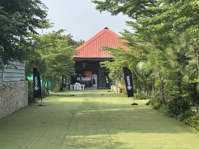 An outdoor pathway lined with trees and BYBIT banners leading to a pavilion with a red roof, featuring an 'IRL Trading' sign.