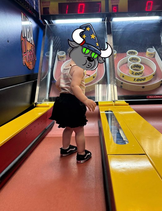 A young girl wearing a cartoon hat with a wizard bull graphic plays a skee-ball arcade game at an arcade.