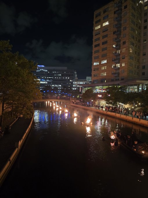 A nighttime cityscape shows a river illuminated with floating candles, reflecting the lights of buildings and a crowd gathered along the walkway.