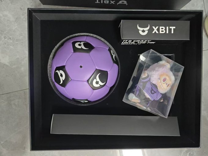 A purple soccer ball and a small plush figure of "Labubu" (a popular character) are displayed in an XBIT-branded gift box, matching the post thanking XBITDEX for the cute Labubu merchandise. The box features the XBIT logo and tagline "IT'S CRAZY UNTIL YOU DO IT."