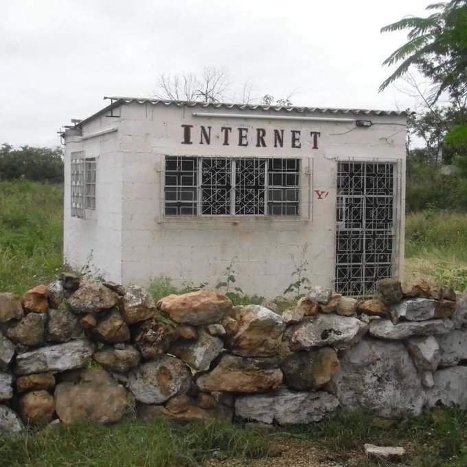 A small white building labeled "INTERNET" with barred windows and door, sitting behind a stone wall in a rural setting.