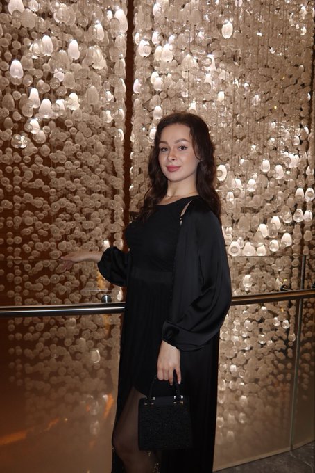 A woman with long dark hair, wearing an elegant black dress with sheer sleeves, holds a small black handbag while posing in front of a stunning wall decorated with cascading seashell-like ornaments. Her sophisticated, professional look complements the post's message about her growing success in the Web 3 industry, reinforcing the celebratory tone of receiving two job offers.