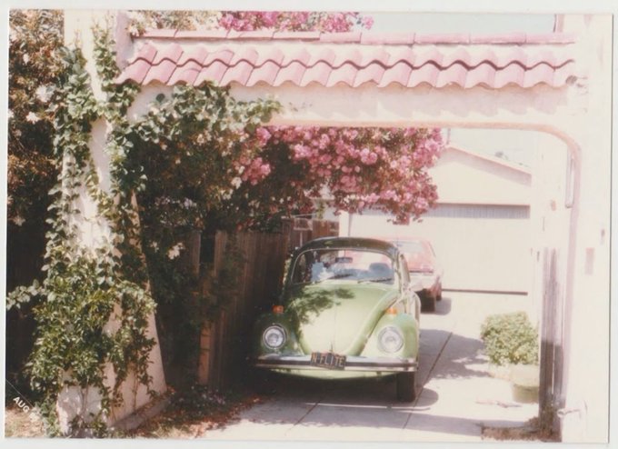 A light green Volkswagen Beetle with a license plate reading "XFL 184" is parked in a driveway under a Spanish-style archway adorned with a red tile roof and lush pink bougainvillea blooms. Ivy climbs the arch's pillar, and another car is partially visible behind the Beetle near a white house.