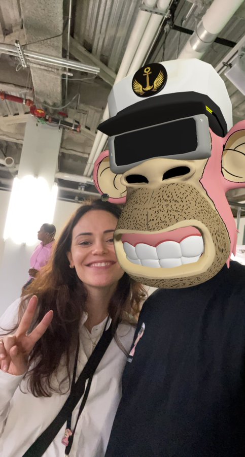 A smiling woman makes a peace sign while posing next to a person wearing a large monkey mascot costume with VR goggles and an anchor cap.
