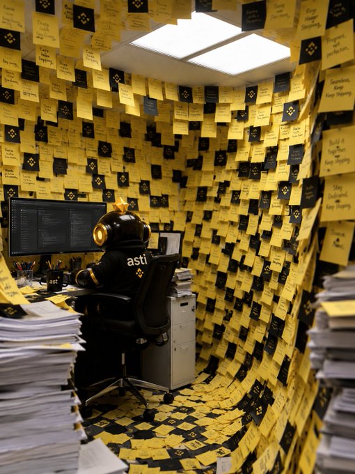 A lone worker in an "asti" branded spacesuit-like helmet sits at a computer terminal, surrounded by thousands of yellow and black sticky notes completely covering the walls, ceiling, and floor of the office space. The "Quietly building what comes next" caption reflects the scene's atmosphere of solitary, focused work amid an overwhelming storm of handwritten tasks and ideas crowding the room.