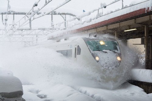 A train pushes through heavy snow at a station platform during a severe snowstorm, with snow blasting in all directions.