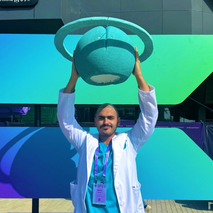 A smiling man in a white coat and teal scrubs with a 'BP25' lanyard lifts a teal spherical trophy above his head outdoors.