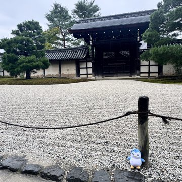 A small cartoon gnome figure stands on a stone border in front of a raked gravel zen garden at a traditional Japanese temple gate.