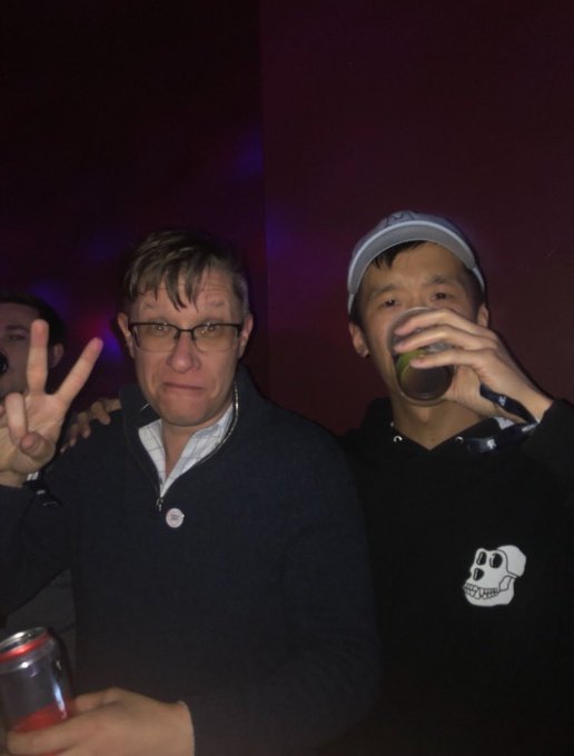 Two men posing for a photo in a dimly lit club setting; the older man on the left makes a peace sign while wearing glasses and a dark sweater, and the younger man on the right drinks from a glass while wearing a black hoodie and a cap.