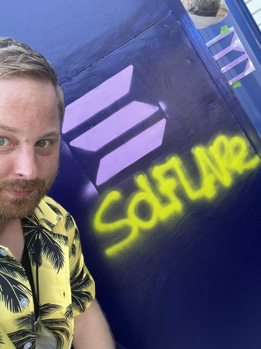 The image features a man wearing a yellow shirt with palm trees printed on it, standing next to a blue wall covered in graffiti. The graffiti includes the word "Solflare" written in yellow letters above the word "Sudflare". The caption of the image reads "Just use Solflare My favorite feature (actually 2): - The Debit Card - Private Send Next that they have a kickass UI/UX experience".