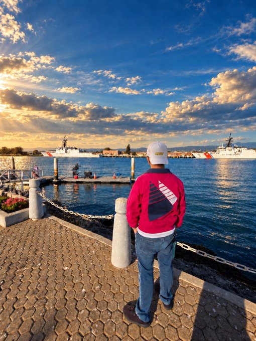 A person wearing a red sweater with a sailboat design, blue jeans, and a white cap stands on a waterfront promenade looking out at a harbor with naval ships at sunset.