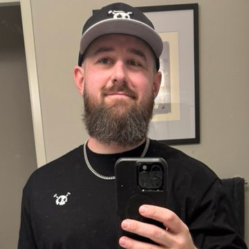 A mirror selfie of a smiling man with a brown beard, dressed in a black t-shirt and a black and grey baseball cap, holding a smartphone.