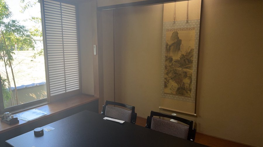 An interior view of a traditional Japanese-style room featuring a dark wooden table with cushioned chairs, a hanging scroll painting on the wall, and a window with slatted blinds revealing outdoor greenery.