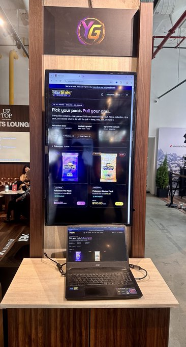 A kiosk featuring the 'YG' logo displays Pokemon trading card packs on a touchscreen, with a laptop showing a similar interface on the counter below.