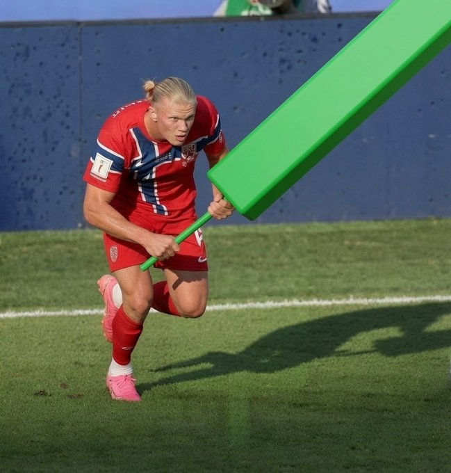 A digitally edited image shows a female soccer player in a red Norway uniform running on a green field while holding a comically oversized bright green stick or bat.