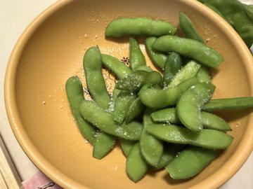 A simple photograph of a tan/yellow bowl filled with salted edamame (green soybean pods) sprinkled with coarse salt, served on what appears to be a restaurant table.