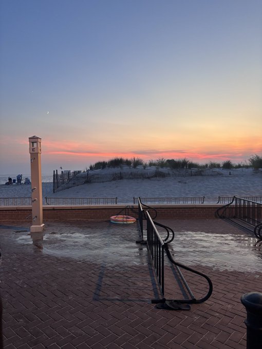 A beachfront boardwalk at sunset with brick paving, metal railings, a shower station, sandy dunes, and a colorful sunset sky.