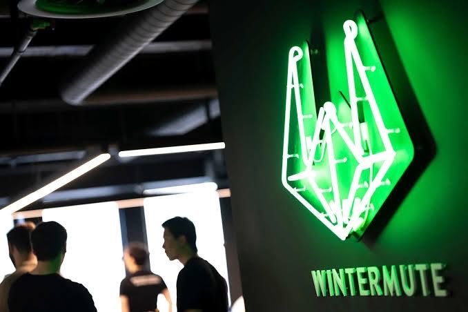 A photograph of a glowing green neon sign displaying a geometric fox logo and the text 'WINTERMUTE', with people visible in the background.