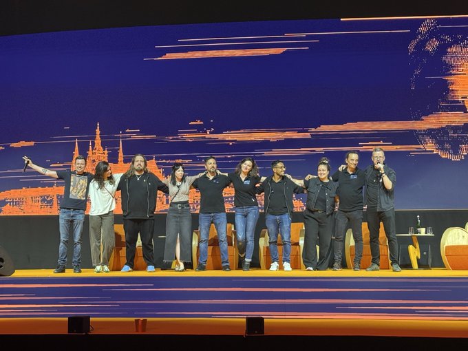 The image features a group of people standing together on stage, posing for a photo with their arms raised. The background shows an orange sky, which contrasts nicely with the black backdrop of the stage. There are at least nine individuals visible in the picture, all dressed similarly and looking towards the camera. The post includes text that reads "This year, Prague did something impossible. A bear market conference was completely packed the whole time. I’ve been here since the first one, and my professional evaluation is that this was the best one thus far".