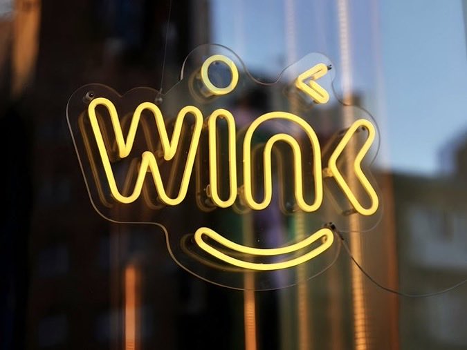 A neon sign glowing yellow in the shape of a smiley face with the word 'Wink' written inside it, hung against a glass window.