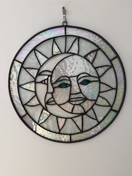 A circular stained glass suncatcher featuring a sun face design with two faces merged together, framed in dark metal with iridescent glass accents, hangs against a white wall.