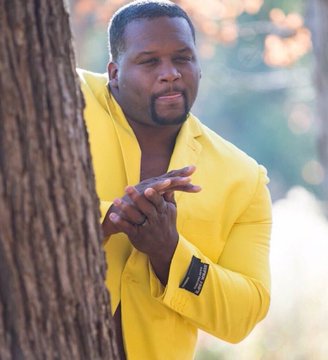 A man in a bright yellow blazer stands behind a tree trunk in a wooded area, gesturing with his hand.
