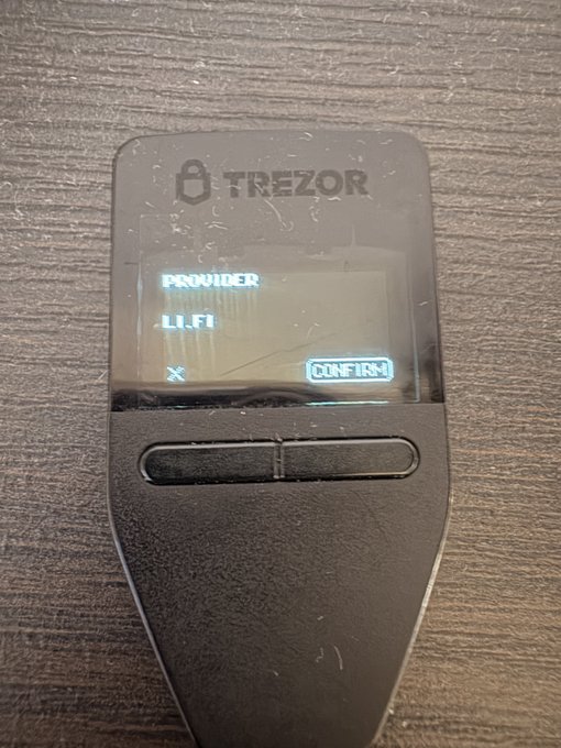 A black Trezor hardware wallet displays 'PROVIDER 1.1.1' on its screen with a CONFIRM button, resting on a wooden surface.