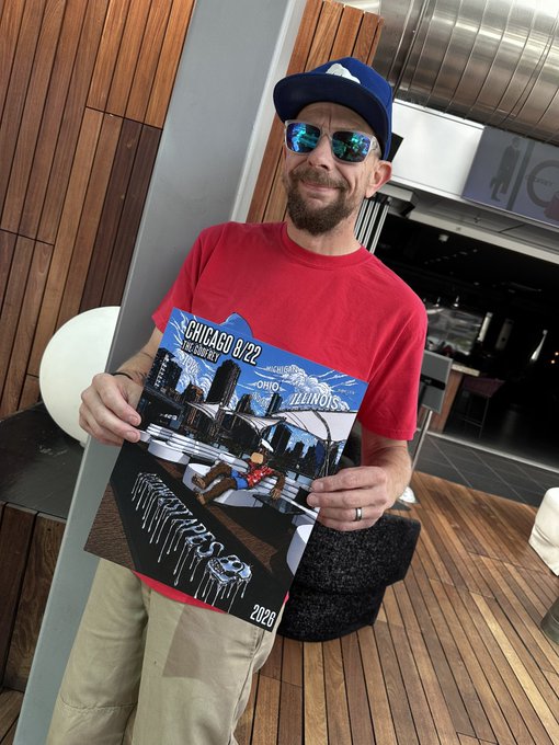 A man wearing a blue cap, sunglasses, and a red t-shirt smiles while holding a Chicago-themed poster for 'The Sixth City', dated 8/22, created in collaboration with 'Runaway Press' and 'Ohio' (2026).