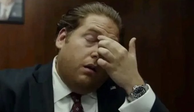A man in a dark suit and tie with a small American flag pin rubs his eyes in apparent frustration while seated at a desk.