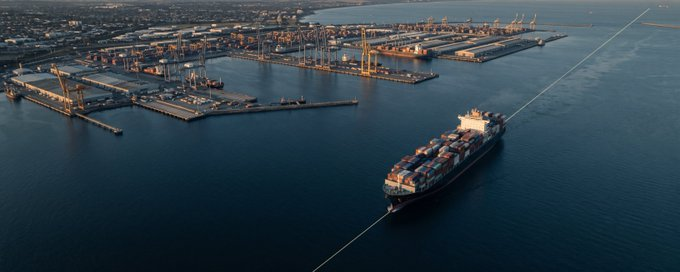 An aerial view of a large container ship sailing out of an industrial port with cranes and shipping facilities visible along the coastline.