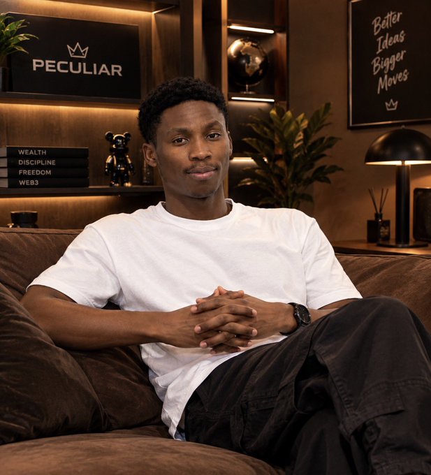 A young man in a white t-shirt sitting on a brown leather couch in a room with decorative elements, including books labeled "WEALTH, DISCIPLINE, FREEDOM, WEB3" and signs reading "PECULIAR" and "Better Ideas Bigger Moves".