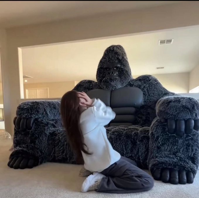 A woman kneels on the floor in front of a massive, oversized chair covered in dark fur and featuring a gorilla head and large fist-shaped armrests.