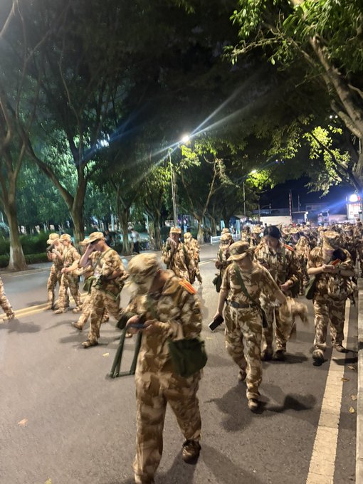 A nighttime photograph captures a large group of soldiers in camouflage uniforms marching down a tree-lined street, carrying gear and rifles.