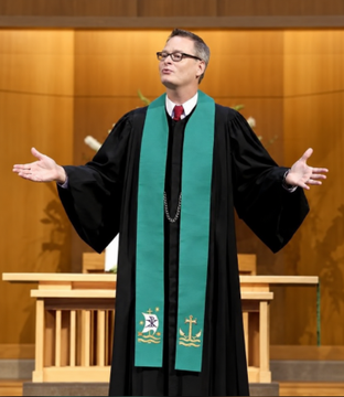 A pastor wearing a green stole over a black robe with arms outstretched in what appears to be a church sanctuary.