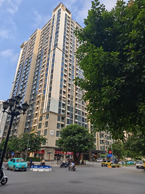 A tall, modern residential apartment building stands at a city intersection, framed by large green trees and streetlamps, with cars and motorbikes passing by on the road below.