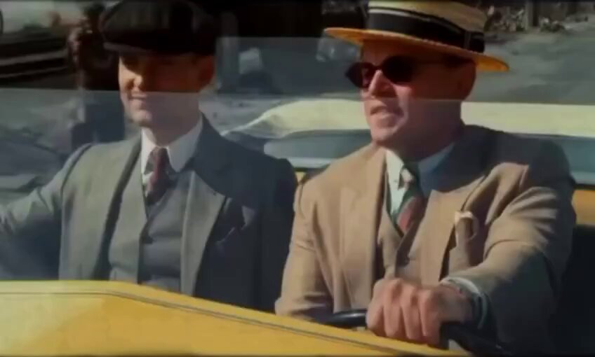The image shows two men sitting inside a car wearing suits and hats. The man on the left side of the car has his arm out, possibly gesturing or pointing at something outside the window. The other man on the right side of the car is looking towards him with interest.