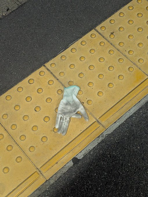 A small, silver-colored sculpture of a hand resting on yellow tactile paving tiles next to a street curb.