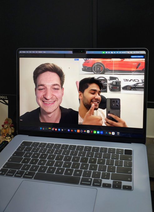 A silver laptop displays a photo of two men smiling, with one holding a smartphone taking a selfie, alongside images of a red sports car in the background.