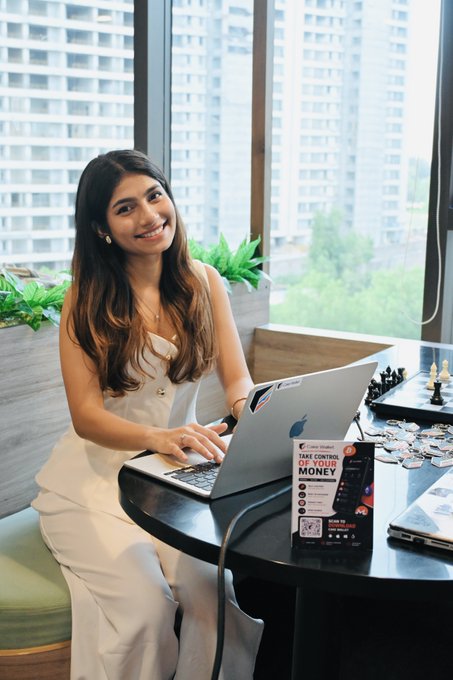 A smiling woman with long dark hair sits at a table with a MacBook laptop, with a promotional card visible that reads "TAKE CONTROL OF YOUR MONEY" with a QR code.