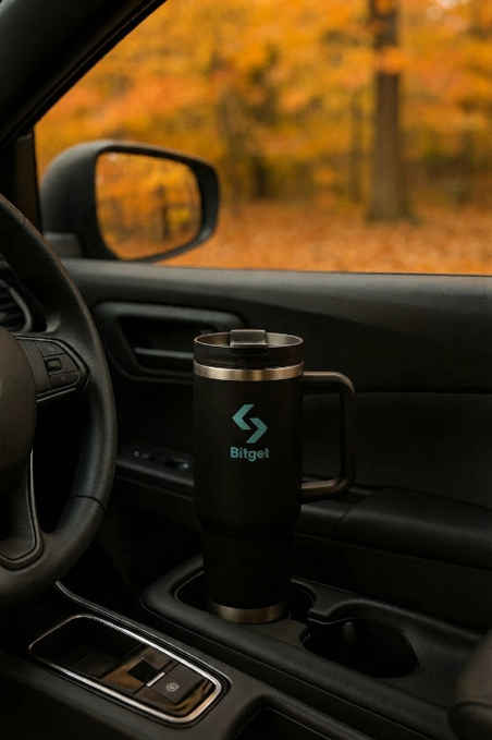 A black insulated travel mug with a handle sits in a car's cup holder, displaying the teal 'Bitget' logo, with autumn foliage visible through the side window.
