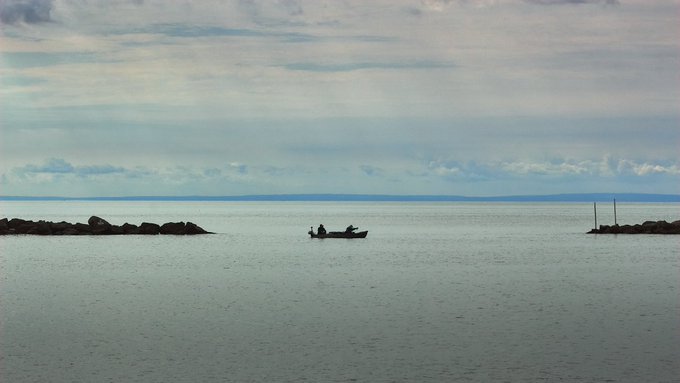 A wide seascape featuring a small boat with two figures floating on calm water, flanked by rocky breakwaters under a cloudy sky.