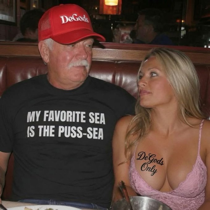 An older man in a red 'DeGods' cap and a t-shirt reading 'MY FAVORITE SEA IS THE PUSS-SEA' sitting next to a blonde woman in a pink top that says 'DeGods Only'.
