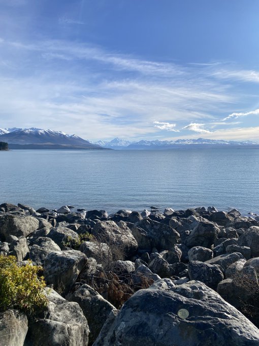 A scenic view of a large blue lake bordered by large gray rocks in the foreground, with snow-capped mountains visible in the distance under a partly cloudy sky.